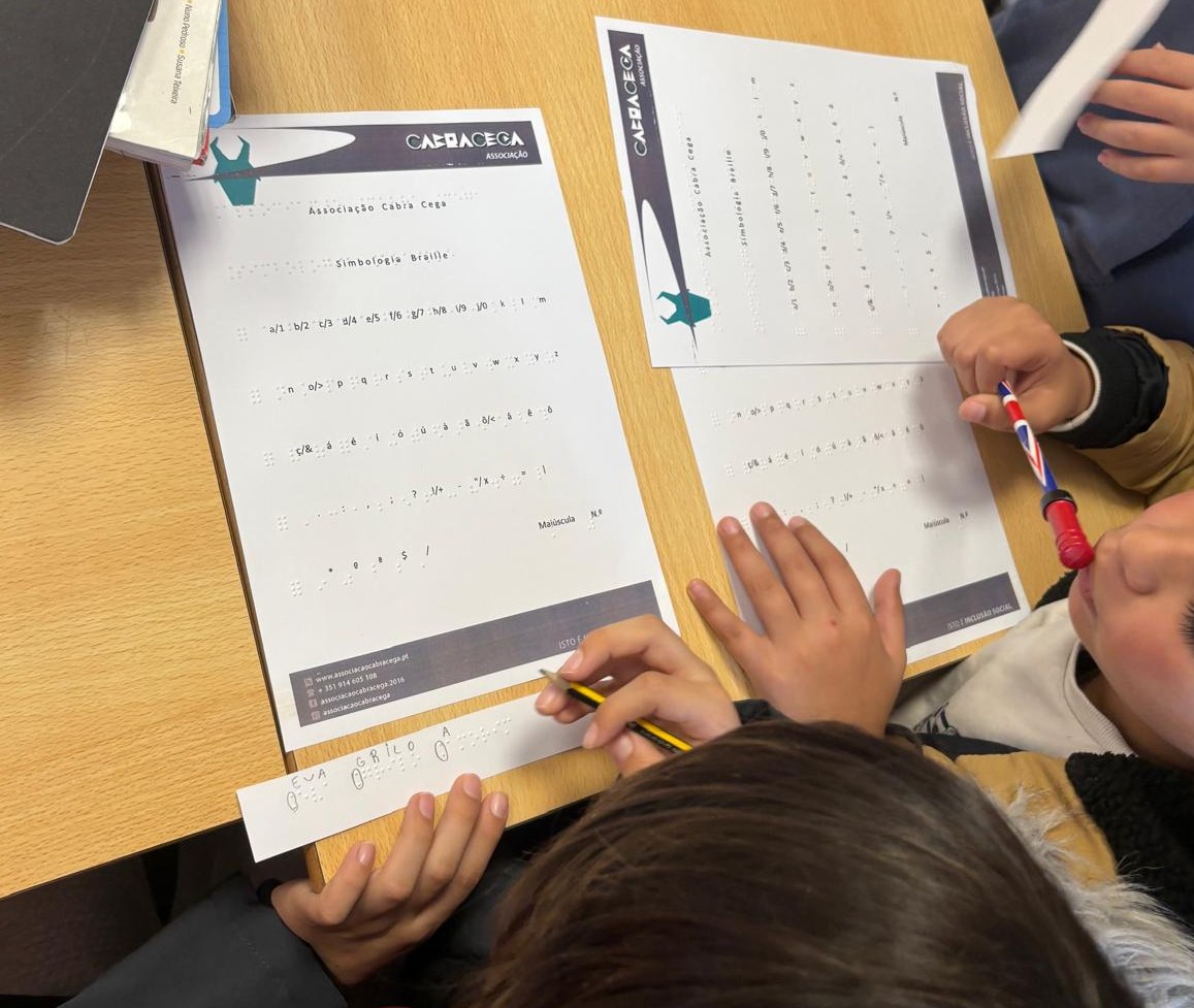 A imagem mostra duas folhas de papel sobre uma mesa de madeira. As folhas têm o logotipo da Associação Cabra Cega no canto superior direito. O título em ambas as folhas é Simbologia Braille. Abaixo do título, há uma série de caracteres em Braille, seguidos por letras e números. Duas crianças estão interagindo com as folhas, uma delas segurando um lápis e a outra uma caneta com o padrão da bandeira do Reino Unido. Uma das crianças está escrevendo em uma tira de papel com caracteres em Braille e letras que formam as palavras EVA e GRILO.